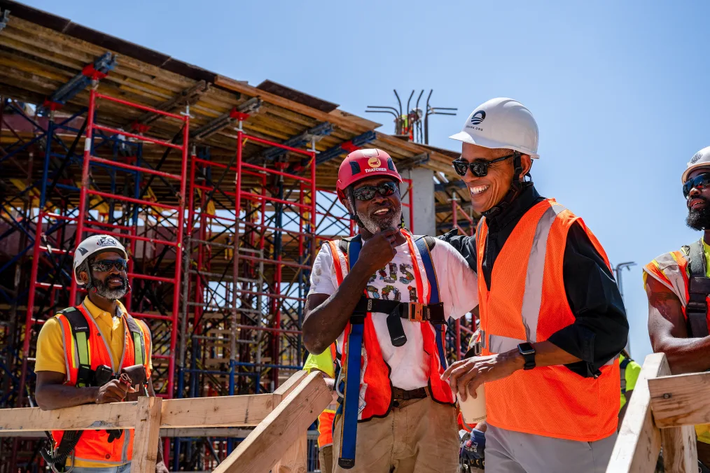 President Obama stands in the middle of the Obama Presidential Center construction site. He is holding a cup of coffee and wearing an orange safety vest and a white hard hat with the Obama Foundation rising sun logo. Behind him are three Black men with deep skin tones. They are smiling and wearing orange safety vests and white and orange hard hats. 
