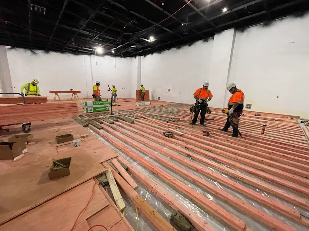 Construction workers wearing bright yellow or orange shirts and hard hats look down at horizontal rows of reddish wooden beams laid out uniform rows. In the distance, other workers walk on plywood boards that sit on top of the beams.