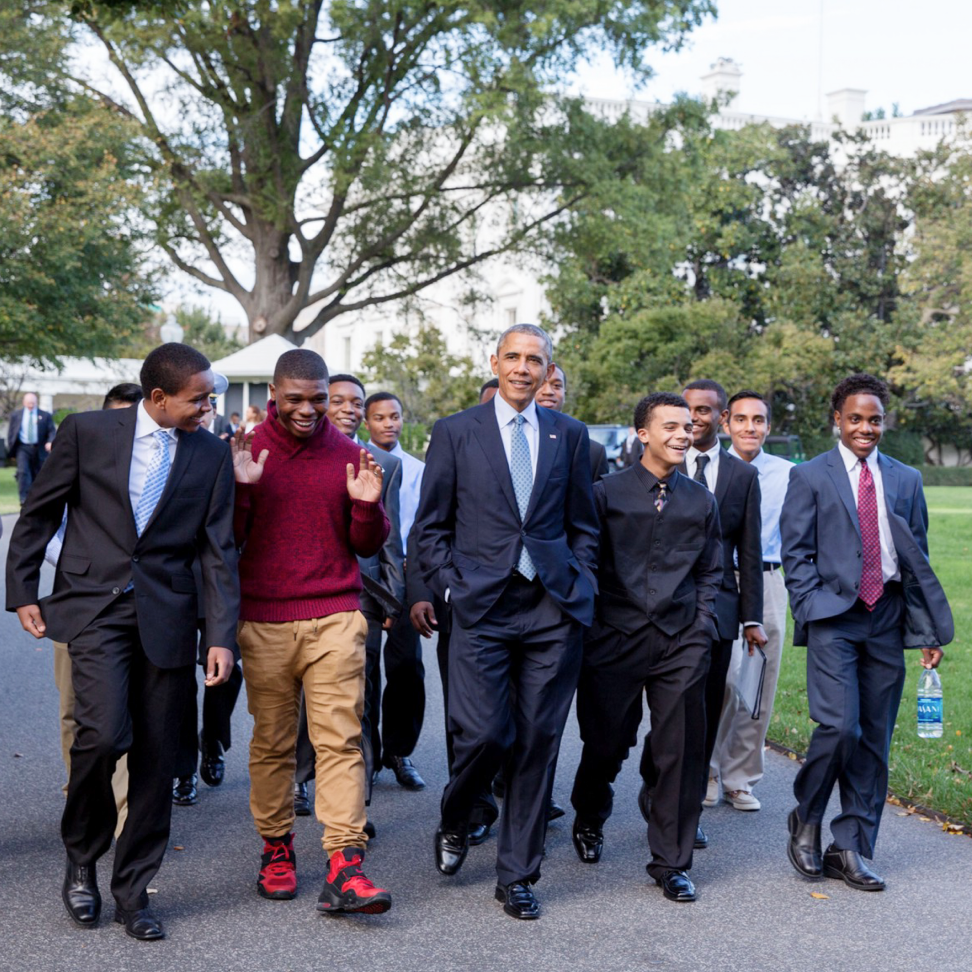 On the South Lawn, President Obama walks in the middle of a group of young men. All have a range of light to deep skin tones. They are wearing a mix of casual and professional attire.