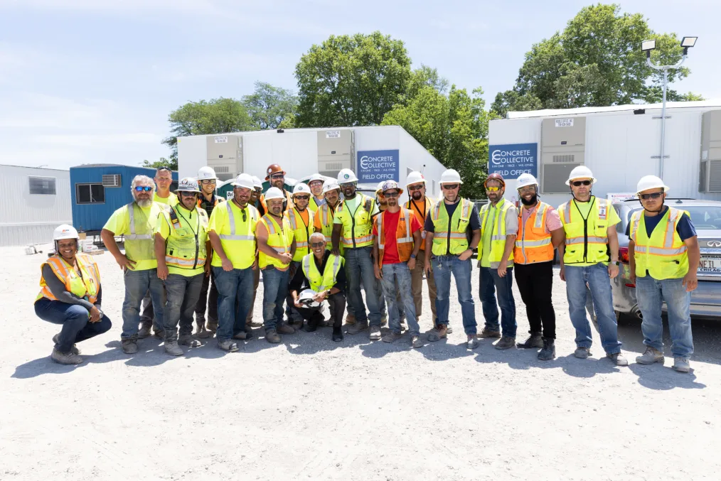 A group of people wearing neon yellow and orange vests and construction hats pose for the camera. Behind them are trailers with signs reading "Concrete Collective Field Office."
