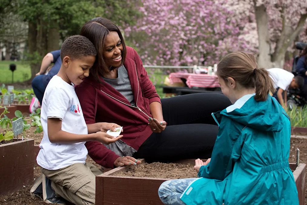 Mrs. Obama plants in the White House Kitchen Garden. She is next to a young Black boy with a medium skin tone and a White girl with a light skin tone and brown hair. All are dressed casually. 