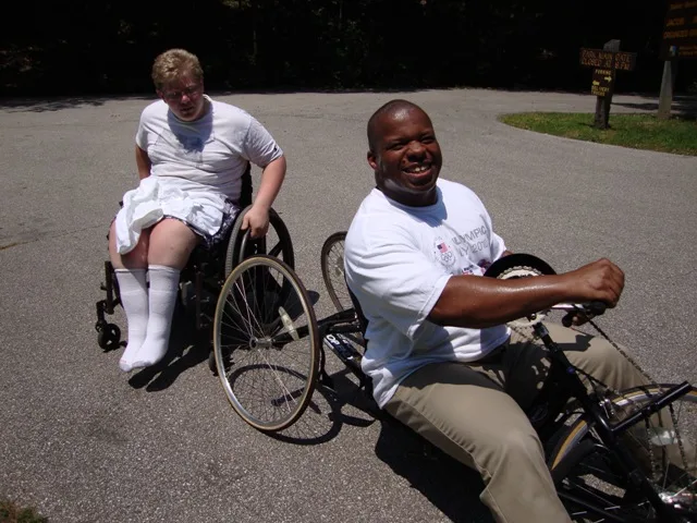 A man with a light skin tone wears glasses and a white shirt and sits in a wheelchair. Beside him is a man with a deep skin tone smiling as he sits in a wheelchair.