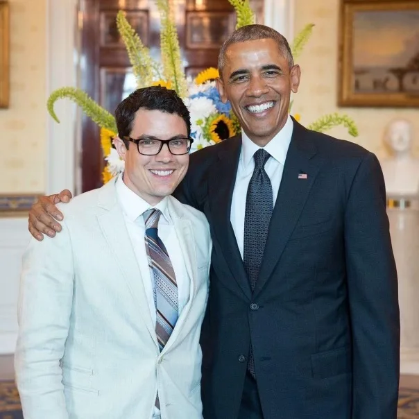 A photo of Dylan Orr posing with President Barak Obama. Dylan has an olive complexion with dark wavy hair, and black glasses. He is wearing a light gray suit with a striped tie. President Obama has a medium deep complexion with closely cut black and gray hair. He is wearing a black suit with a black striped tie. Both Dylan and President Obama are smiling in the photo.