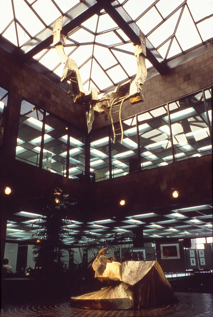 A two-piece, abstract sculpture shown in a massive lobby that features a skylight casting shadows upon its shape.