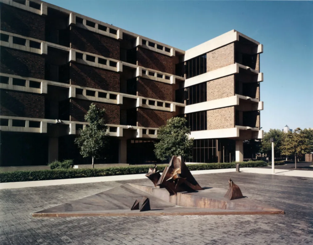 An abstract sculpture installed in a recessed area within a courtyard. In the background, a large building can be seen.