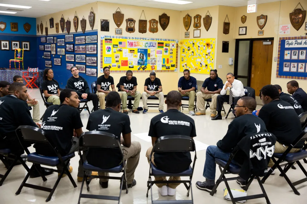President Obama sitting in a circle group of medium to deep skin toned younger to middle aged men in a Hyde Park Academy classroom. President Obama wears business casual attie and the men are wearing black BAM youth guidance tshirts and kahkis. The group is meditating.