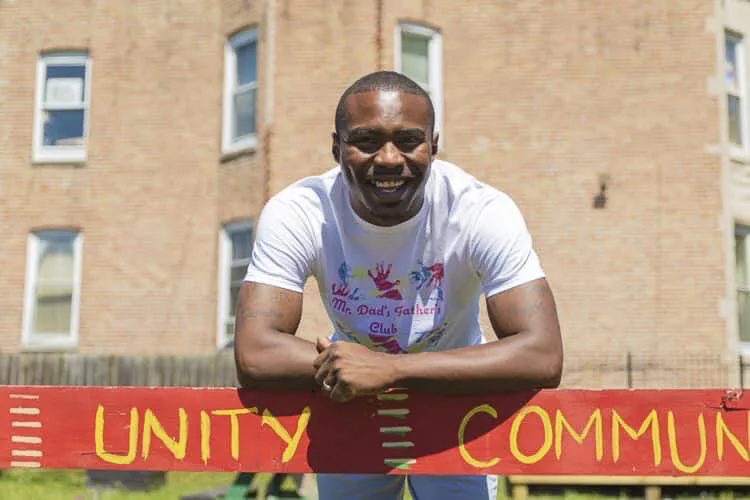 A man with a deep skin tone smiles as he stands over a red sign with yellow lettering that says, "UNITY" and "COMMUNITY" outside of an apartment building. He is wearing a white shirt that reads, "Mr. Dad's Father's Club." 