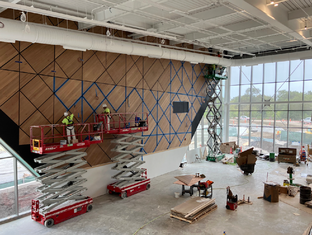 An image of construction being done on Main Court, the Obama Presidential Center's basketball court, at Home Court. The wood panels are on one wall and are brown with blue borders. Each panel of wood is a triangular or diamond shape. Next to the wood panel wall is a glass window. 