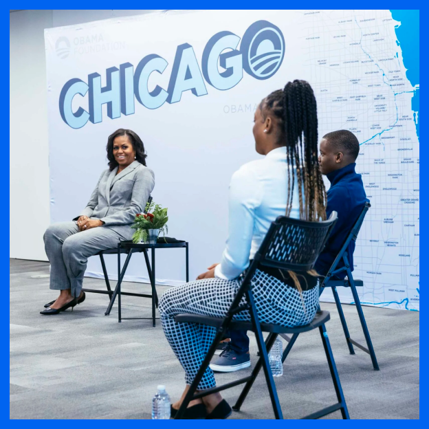 Michelle Obama smiles during a circle group meeting with young men and women. They are evenly spaced out and seated wearing formal attire. In the background, there is a graphic on a wall of the word Chicago and a mini map of the city.