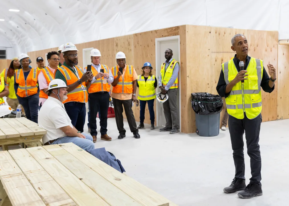 President Obama wearing a black button-up shirt, black pants, black shoes, and a neon vest and holding a microphone speaks to a group of construction workers with a variety of skin tones in a white construction tent. 