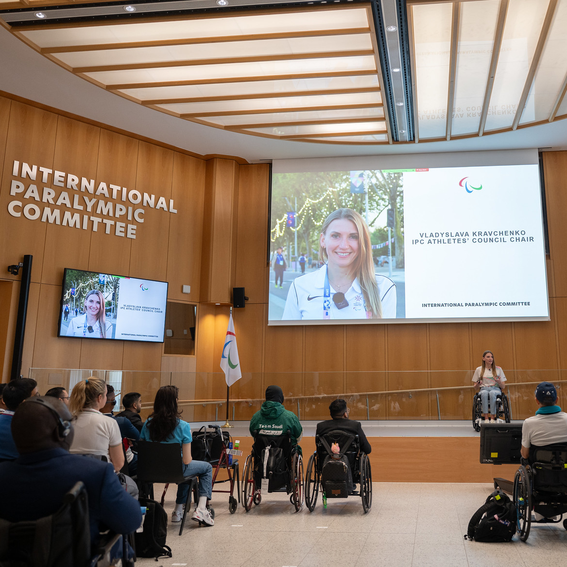 Vladyslava Kravchenko, a woman with a light skin tone and blonde hair, sits in a wheelchair as she speaks on a stage to an audience. A monitor with her face on it is behind her. Words on the auditorium wall read, “International Paralympics Committee.”