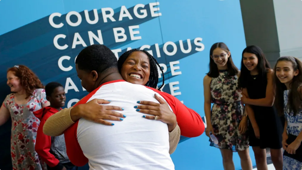 Keke Palmer smiles and hugs a young man in front of a blue wall with an Obama quote on it.