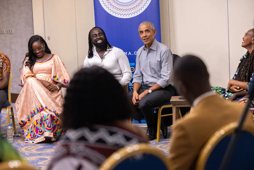 President Obama sits in a room with the members of the Obama Leaders Africa cohort. They are wearing bright colors and are of varying skin tones. The Obama Leaders Africa medallion is in the background.