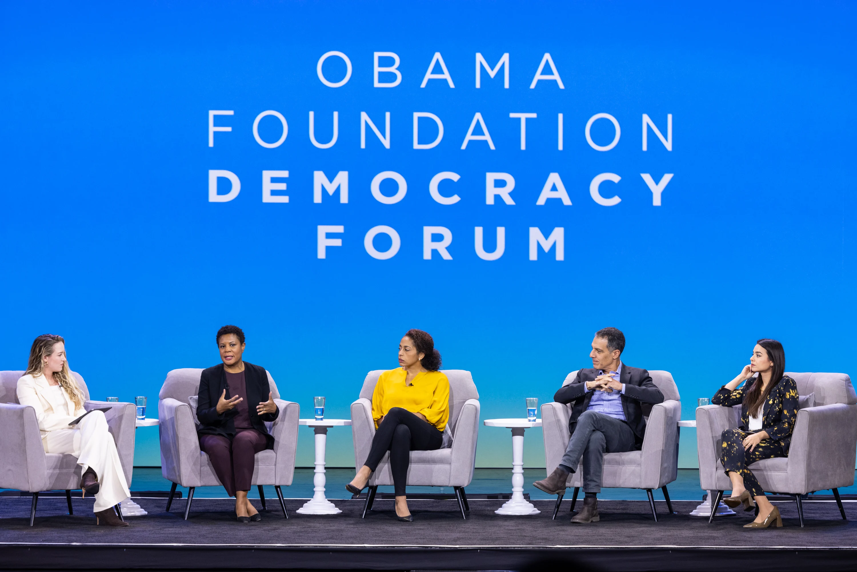 Alondra Nelson, a Black woman with a deep skin tone speaks on a panel alongside  Anna Makanju, Hany Farid, Raquel Vazquez Llorente, and Terah Lyons. All speakers are a range of light to deep skin tones and they sit on a stage. A screen behind them reads, “Obama Foundation Democracy Forum.”