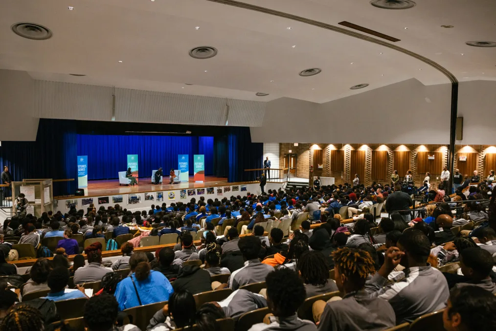 Young people fill a large auditorium. Mrs. Obama sits in a light gray chair next to two young women, also seated.