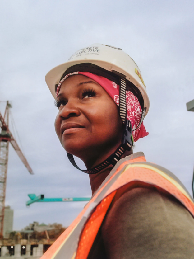 A woman with a medium-deep skin tone wearing an orange, gray, and yellow construction jacket and helmet looks up toward the sky while on a construction site. There are cranes in the background. 