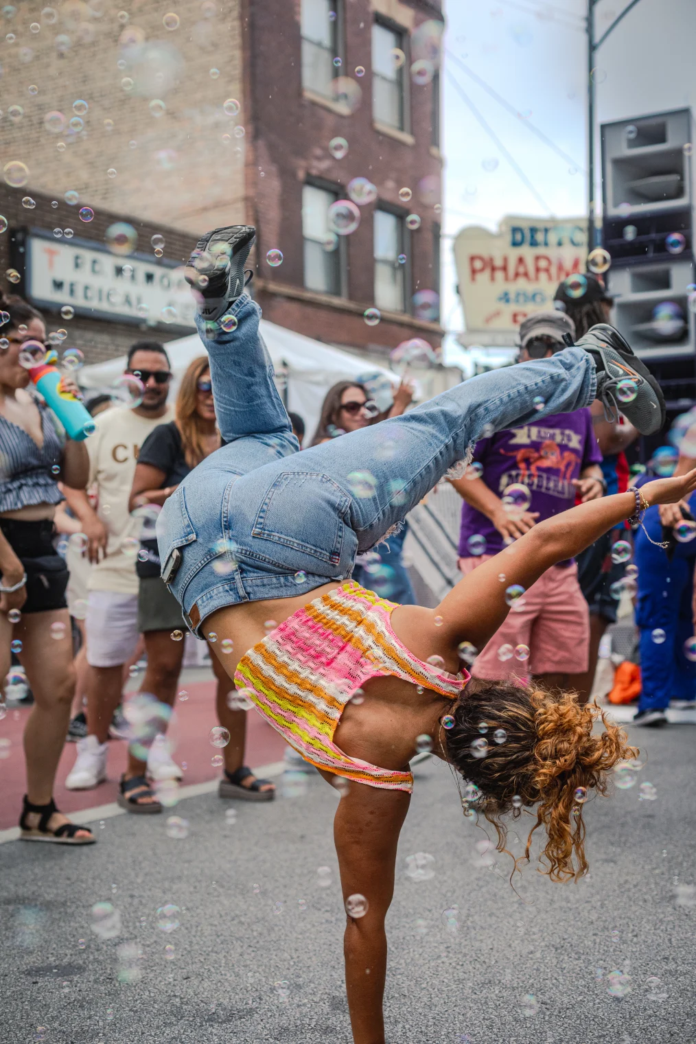 The image is an in motion picture of a person break dancing on the street. The image is outside on a city street. In the background is a brick wall with a signs on it. In the picture are several people, surrounding the breakdancer, as they dance on the street. The breakdancer, who is in the center of the picture, is standing on one hand with their feet in the air. The dancer has on a orange, pink, yellow, and white sleeveless crop top, blue jeans, and grey sneakers. Around the picture are bubbles throughout the air.