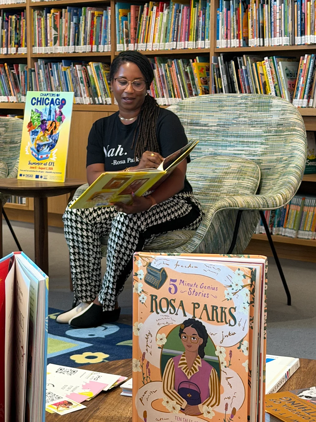 A shot of Ebony Wilkins, A Black woman with a dark skin tone and box braids, reads her book to a group of children with a range of light to dark skin tones at the Chicago Public Library Branch at the Obama Presidential Center.
