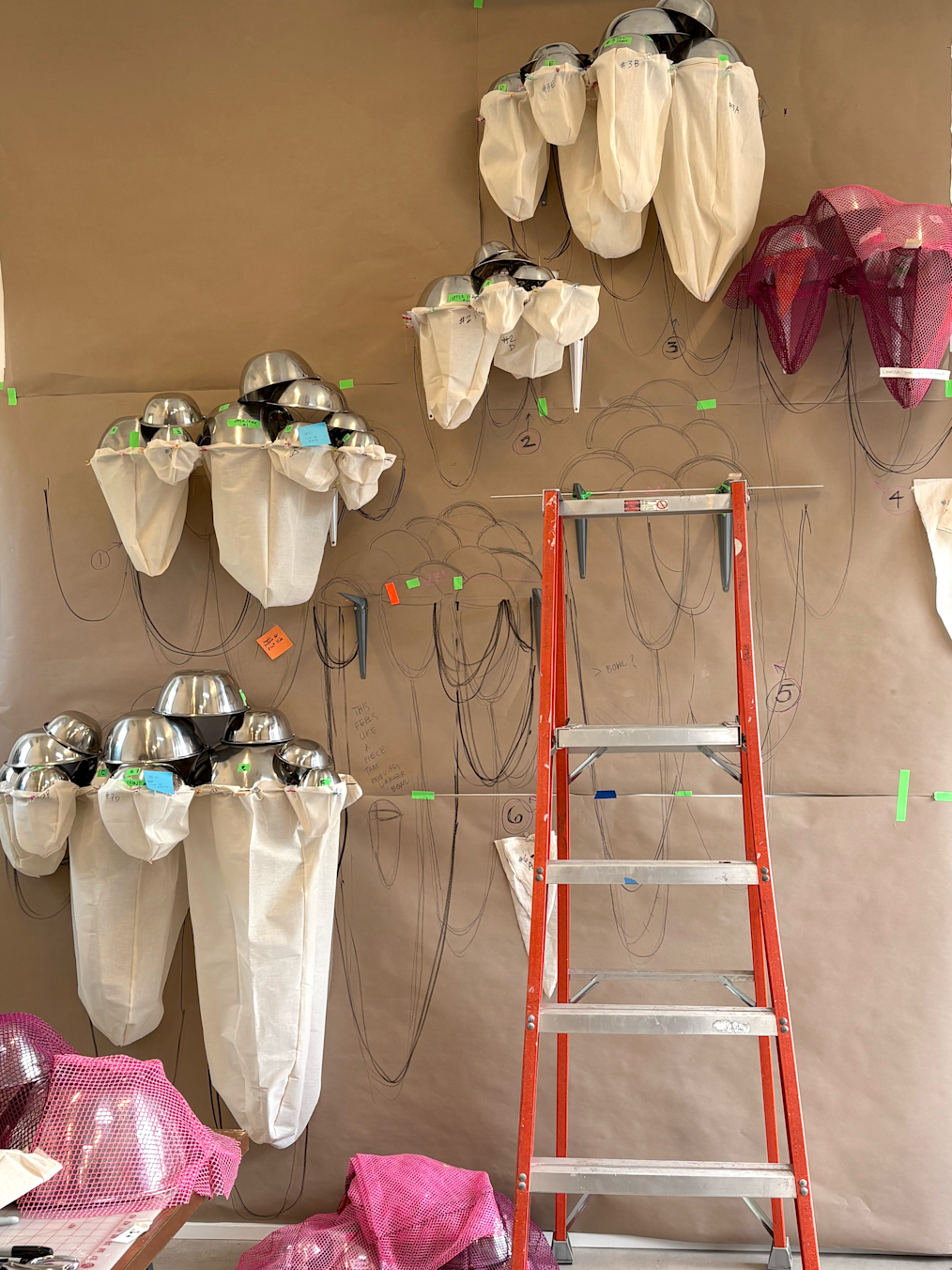 A image shot in Marie Watt's studio that shows the work in progress installation,  “This Land, Shared Sky." The picture is of a wall covered by cardboard paper with writing on it. On the wall are silver bowls combined together in separate clusters. Underneath the bowls in each cluster are white sacks that hold the jingles on the installation. In front of the wall is an orange four step ladder. On the floor of the studio is pink mesh netting. In the left side of the image is a work table. On the work table is a silver bowl upside down and covered with pink mesh netting.