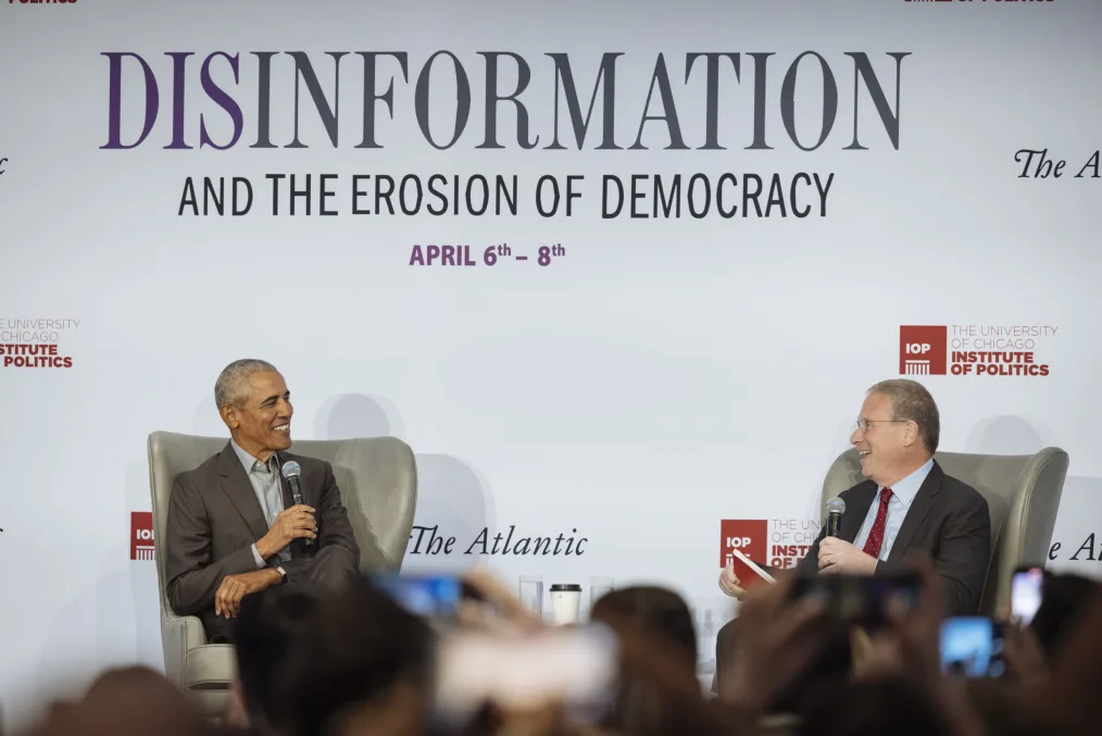 President Obama wearing a dark gray blazer and gray button-up shirt sits on stage in a gray chair with a microphone speaking to a man with a light skin tone wearing a black blazer, a light blue button-up shirt, and a red tie also sitting in a gray chair. In the background there is a light blue backdrop that says "Disinformation" in large letters. 
