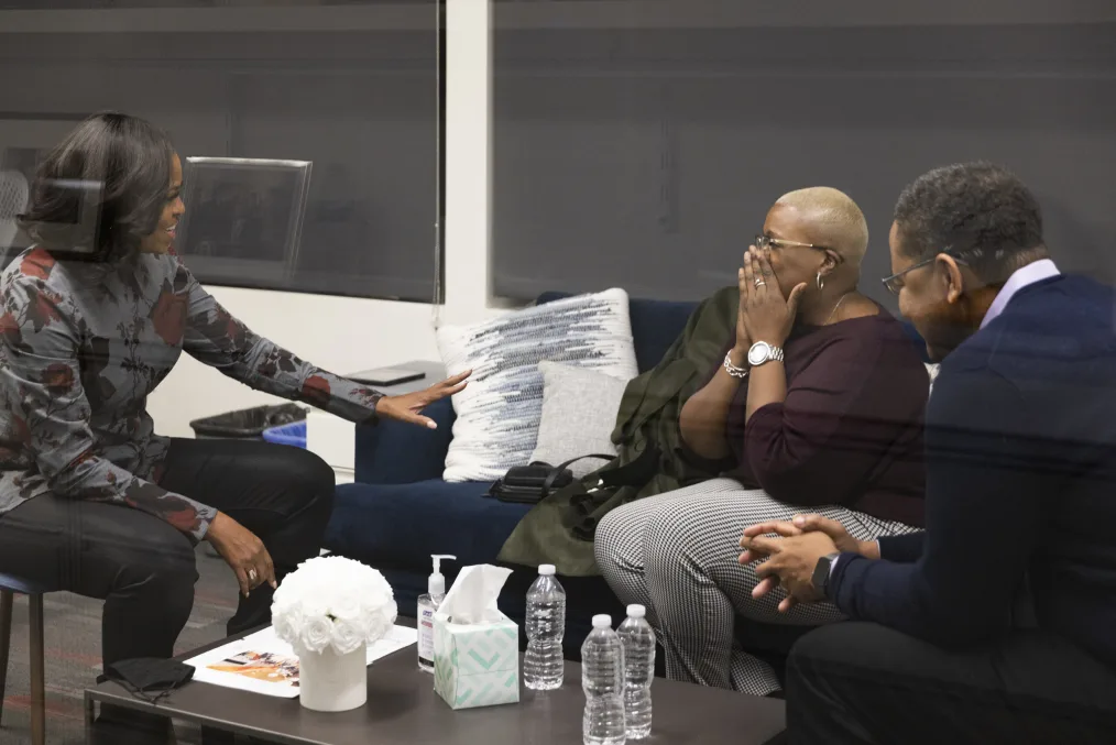 First Lady Michelle Obama wearing a gray sweater with red roses and black pants reaches across from her to a woman with a medium-deep skin tone wearing a burgundy off-the-shoulder sweater, glasses, and black and white plaid pants that is holding her hands up to her face. Sitting next to the woman is a man with a medium-deep skin tone wearing glasses, a navy blue sweater, and black pants.  