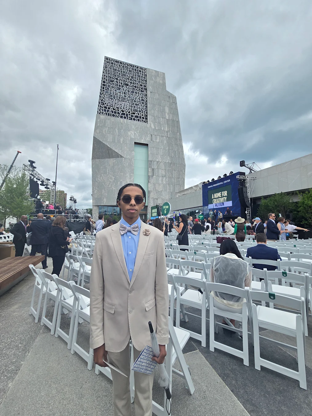 Jonathan Sullen, a 16-year-old Black boy with a light skin tone, smiles as he stands in front of white chairs at the Obama Presidential Center grand opening ceremony. He is wearing a tan suit and a blue shirt. The Obama Foundation Museum Building is in the background. People of a range of light and dark skin tones are in the background. 