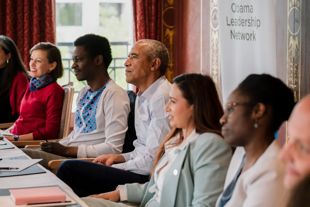 President Obama listens as he sits in between six people with a range of light to deep skin tones. A sign in the background reads, “Obama Leadership Network.” 