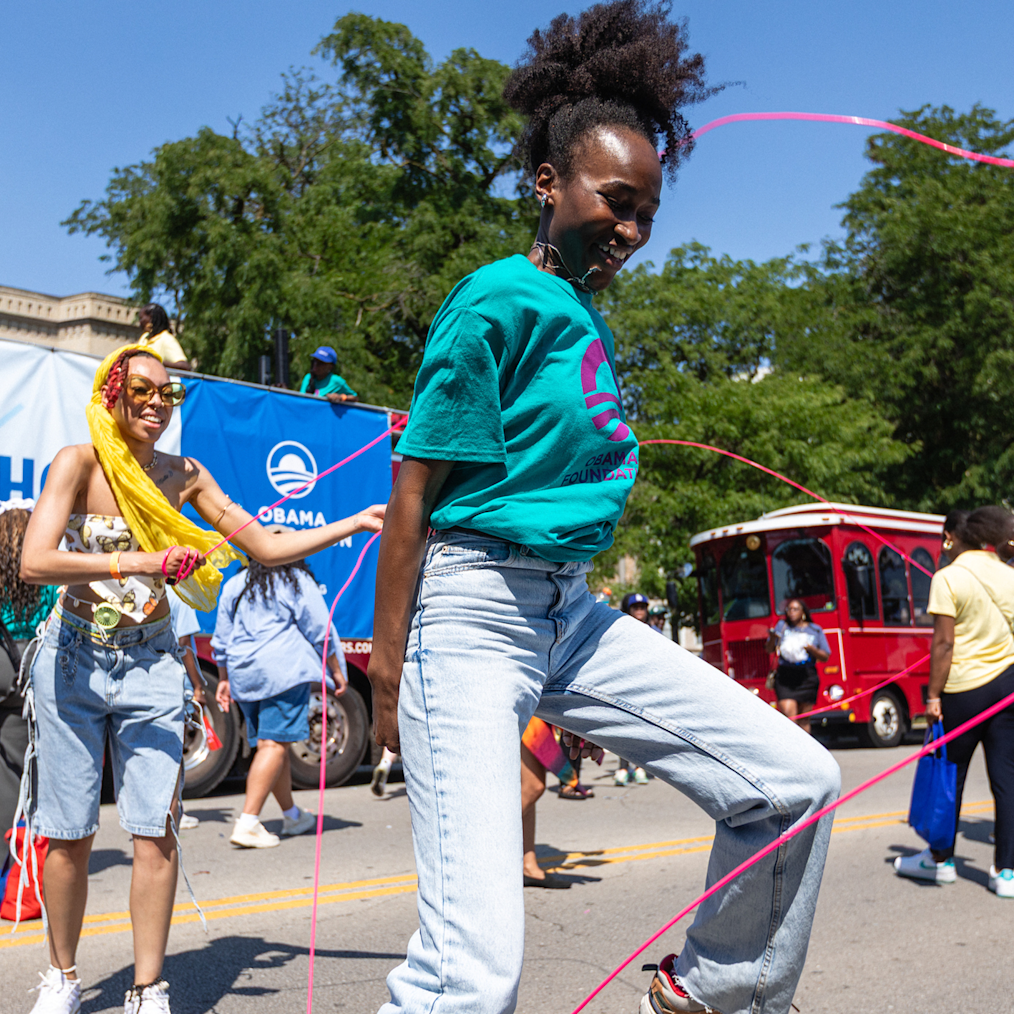 A picture of a young woman with a deep skin tone playing double dutch outside on the sidewalk. She has on a blue and purple Obama Foundation t-shirt.  