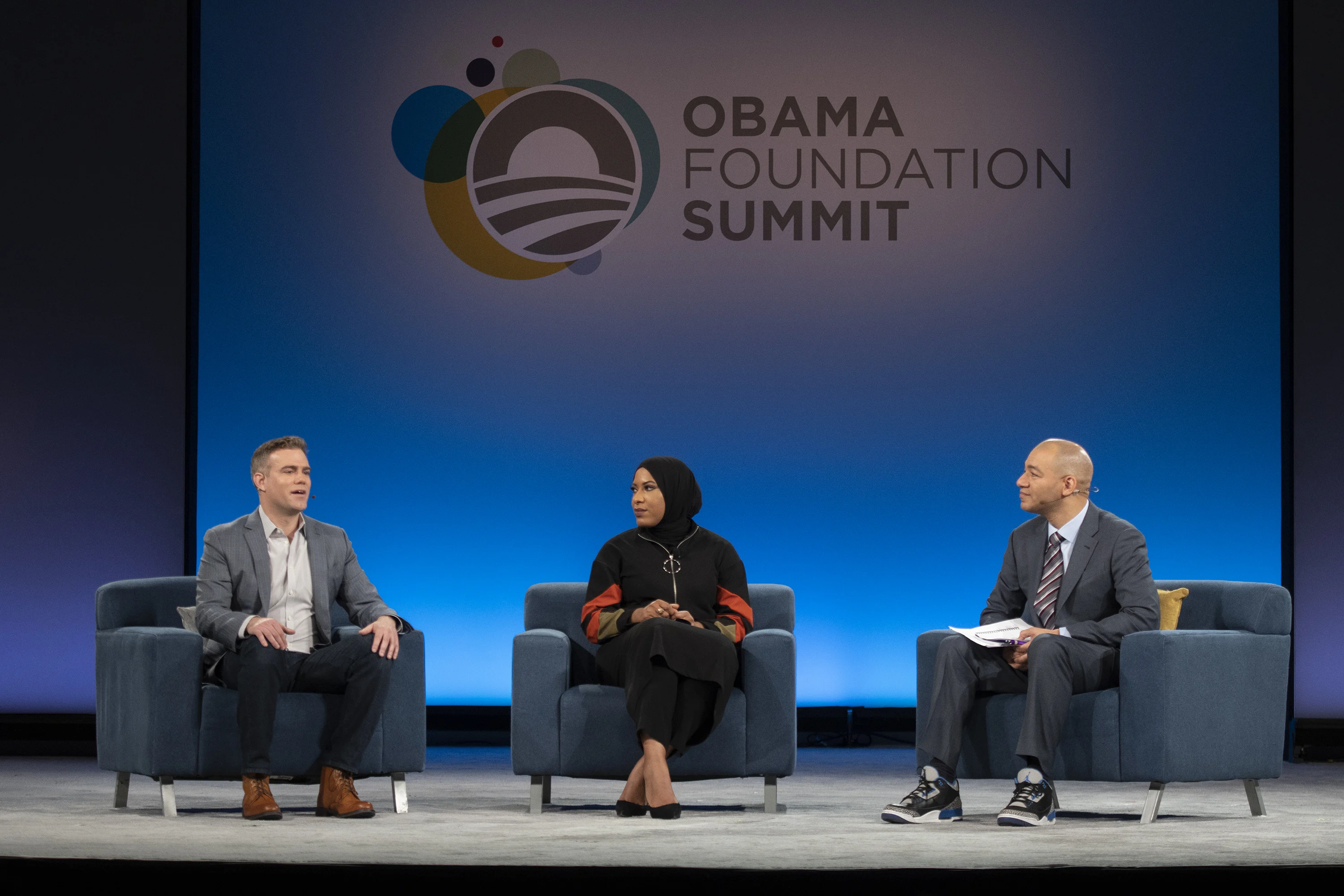 Three people sit in plush chairs on stage. Two men sit on either side of a woman. Behind them is a screen with the words "Obama Foundation Summit"