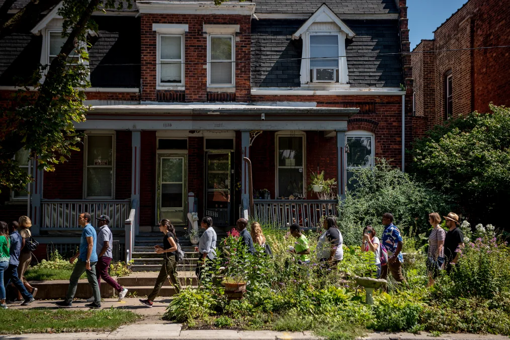 A line of a of tour group walks down the side walk next to a neighboorhood of ok condition, brick houses. The side walk on the right are shrouded by tall plants.