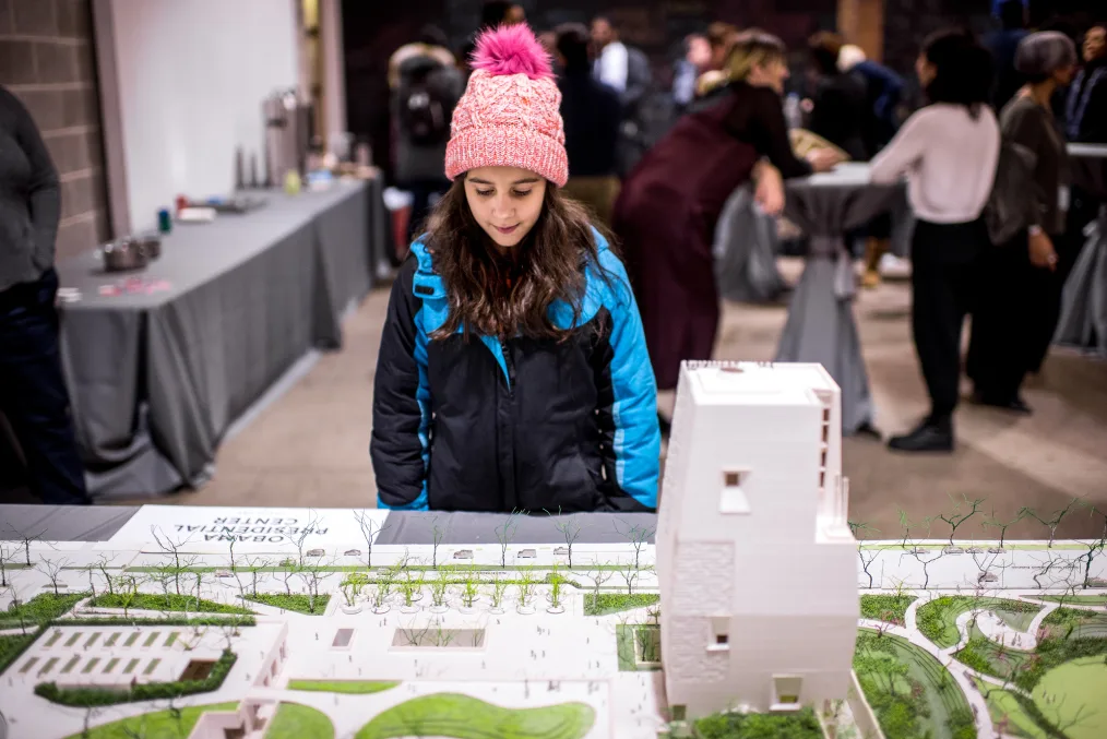 A little girl wearing a pink hat looks down at a model of the Obama Presidential Center. 