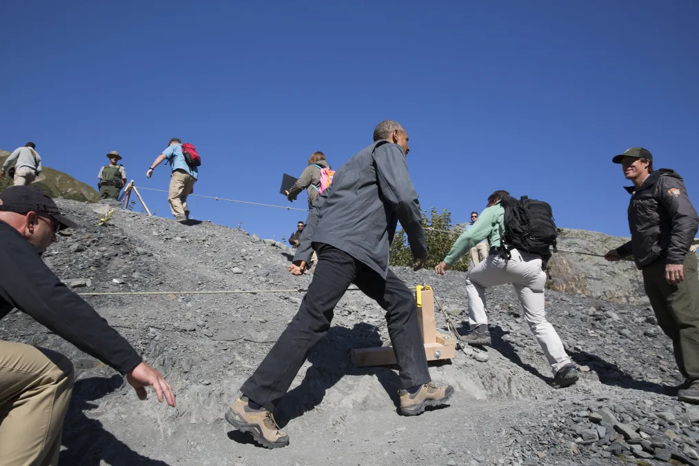 Barack Obama wearing black pants and hiking boots walks uphill along a gray, rocky path outlined by ropes. Others walk ahead and behind him. The sky is a deep blue.
