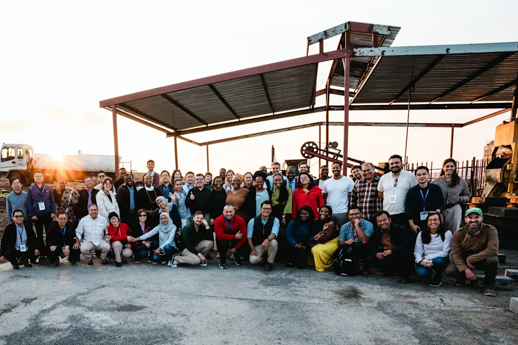 A large group of individuals with a range of light to dark skin tones pose for a photo at sunset. All stand under a canopy. 
