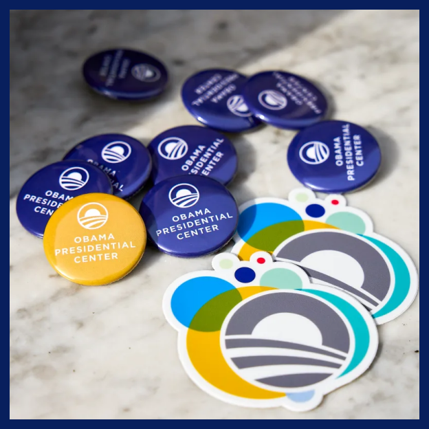 Several blue and yellow buttons that read 'OBAMA PRESIDENTIAL CENTER' and two Obama Foundation logo stickers lay across a sunny table.