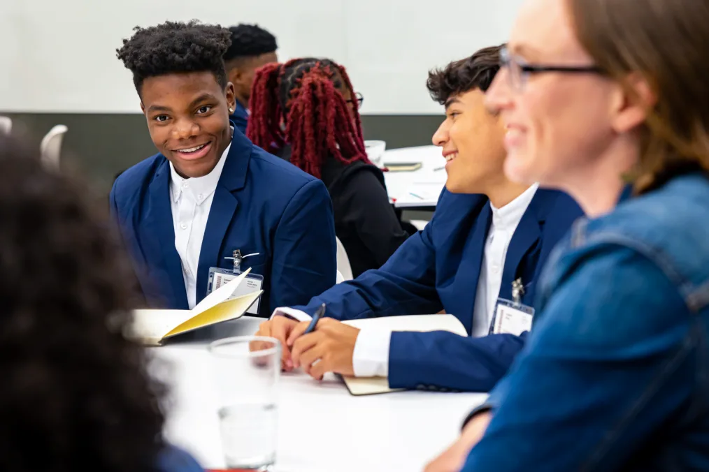 Two young men, one with a medium-deep skin tone the other with a medium-light skin tone sit at a table with a woman with a light skin tone. They all smile and wear business attire.