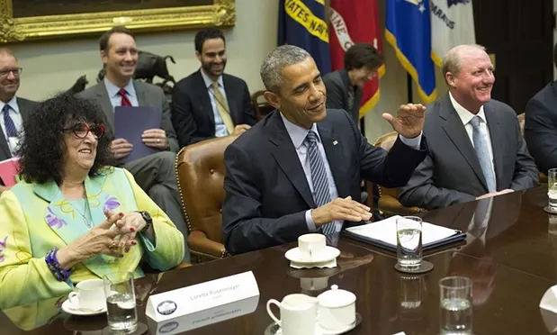 
Alt text: President Obama sits at a table with two others with a light skin tone. Robert Diamond sits behind him alongside two other males with light skin tones. All are dressed professionally. 
