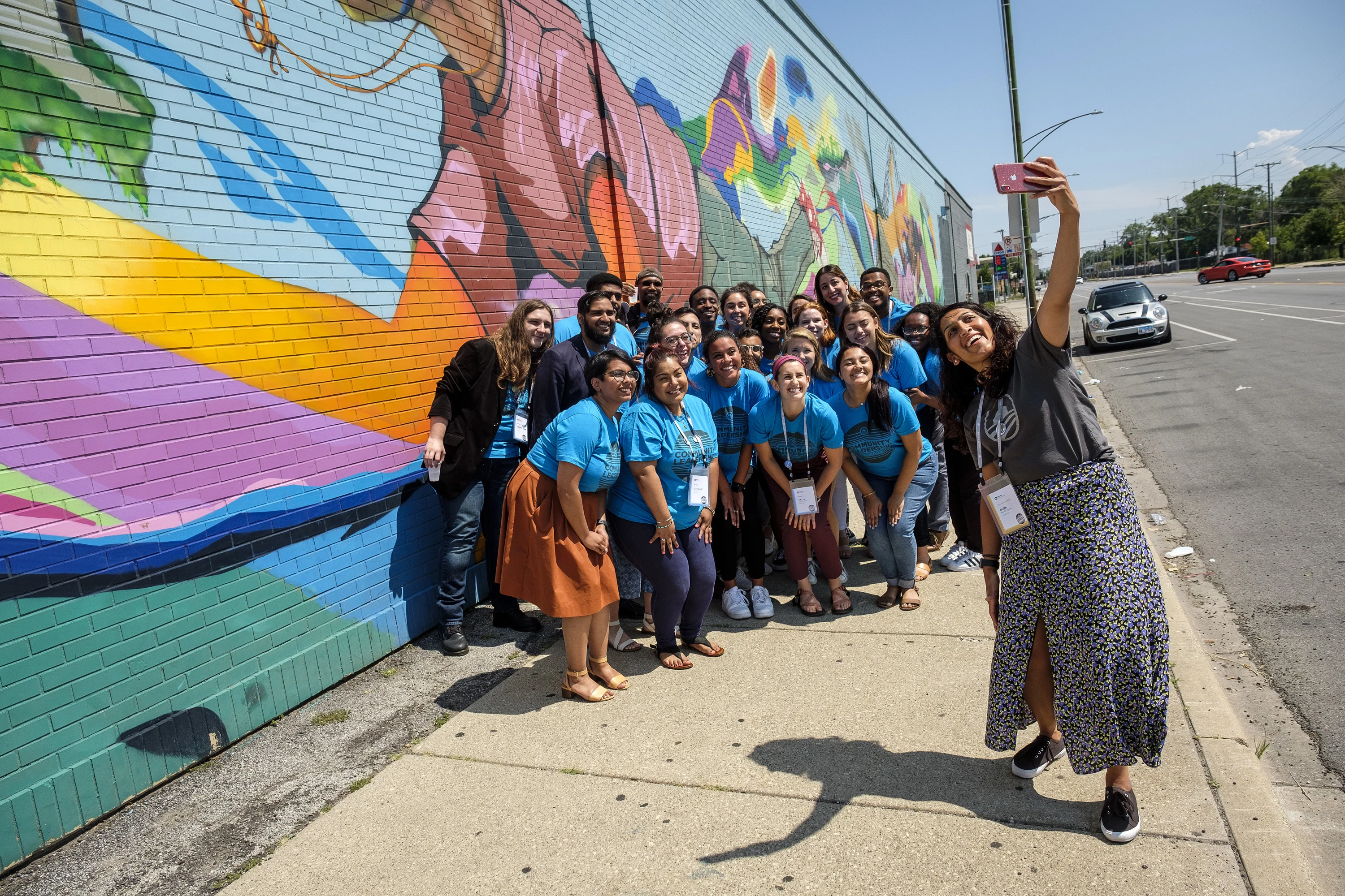 A woman with a medium-deep skin tone, a dark gray Obama Foundation shirt, and a blue, green, black, and pink patterned skirt holds her arm up high with a red iPhone in her hand and stands on a sidewalk, close to the street. There is a group of individuals with a variety of skin tones wearing blue shirts with black letters that spell "Community Leadership" , standing behind the woman. They are all standing against a wall with colorful graffiti art of a person with a medium-deep skin tone and other images. 