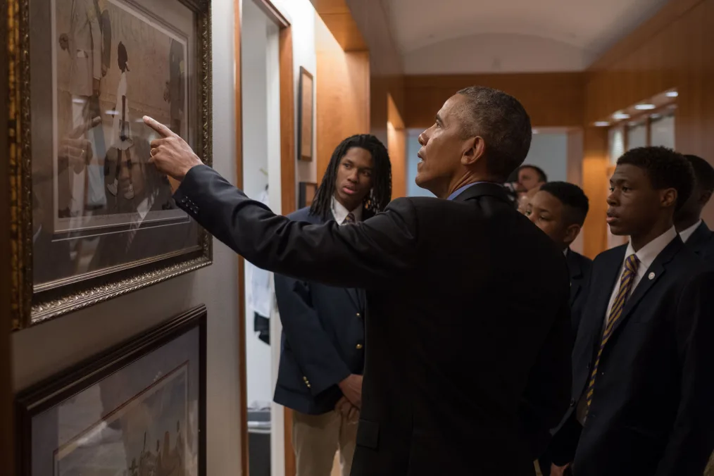 Barack Obama, dressed in a dark suit jacket, is turned away from the camera using his left hand to point to a painting on the wall in a hallway. Four young black men dressed in dark suit jackets and brown pants surround him on his right looking at the painting.