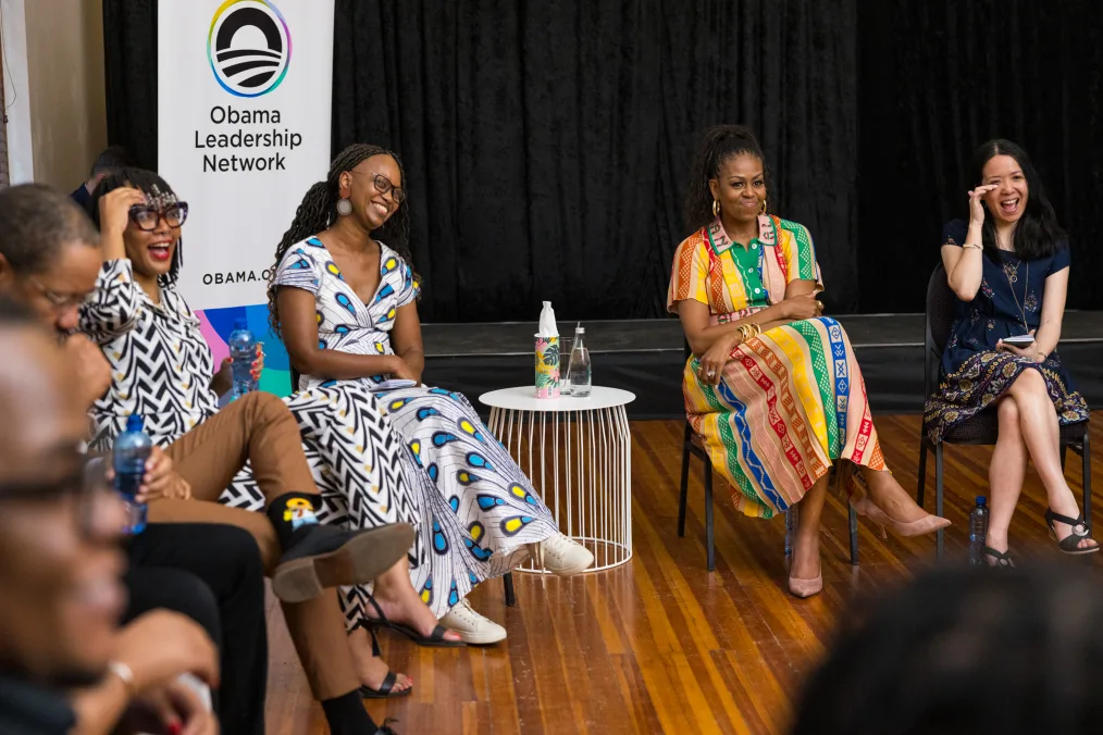 Michelle Obama sits in a circle with women with a range of light to deep skin tones and Michael Strautmanis. Mrs. Obama is wearing a multicolored dress and sits with her legs crossed.  A sign in the background reads, “Obama Leadership Network.” 