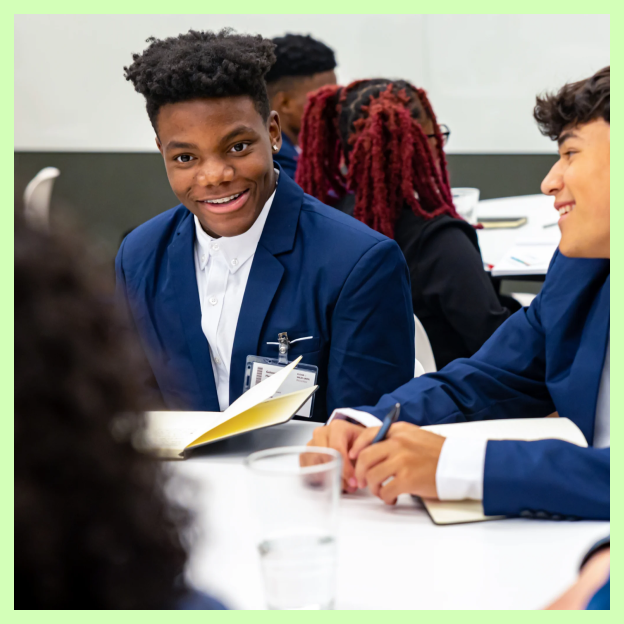 Two young men, one with a medium-deep skin tone the other with a medium-light skin tone sit at a table with a woman with a light skin tone. They all smile and wear business attire.