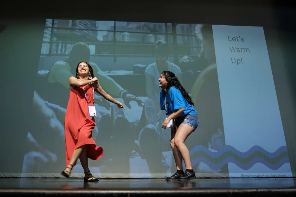 A woman with a light-medium skin tone and a red dress with black flats stands on stage and laughs as she swings her arms and shifts to the left. On stage with her is another woman with a light-medium skin tone, a blue shirt, and denim shorts with black flat buckled shoes, who is also laughing and slightly bent over. They are both standing in front of a screening that says "Let's Warm Up" on the left-hand side with a photo of a few individuals stretching on the right-hand side.  