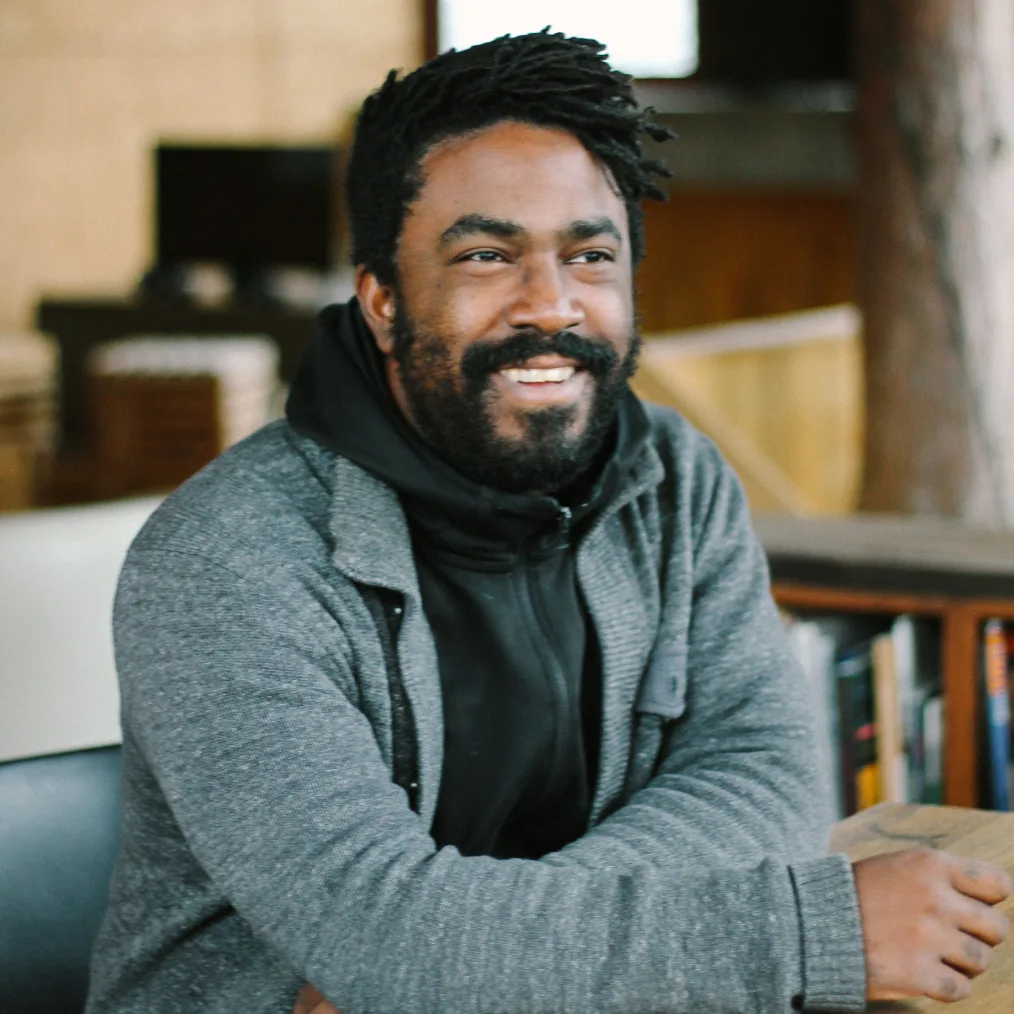 A Black man with short dreadlocks and full beard smiles as he looks off to the right-hand side. His arms are crossed over each other as he sites at a table. Behind him there is a computer and bookshelf. He wears a black hoodie under a gray knit sweater. 
