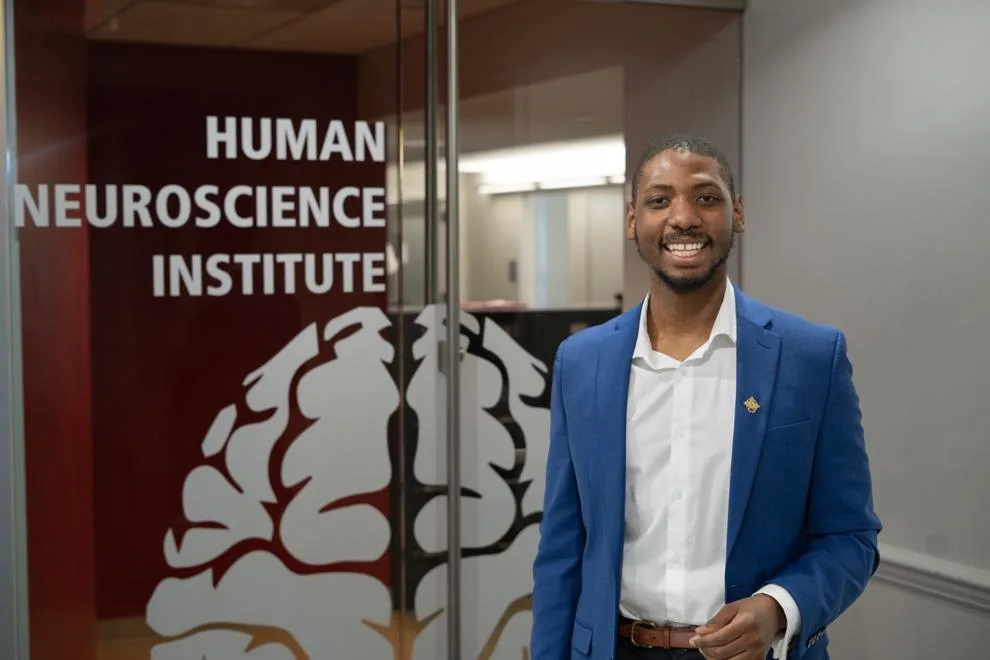 A young Black man wearing a blue suit jacket smiles as he stands in front of glass doors with the words "Human Neuroscience Institute" and brain graphic on them.