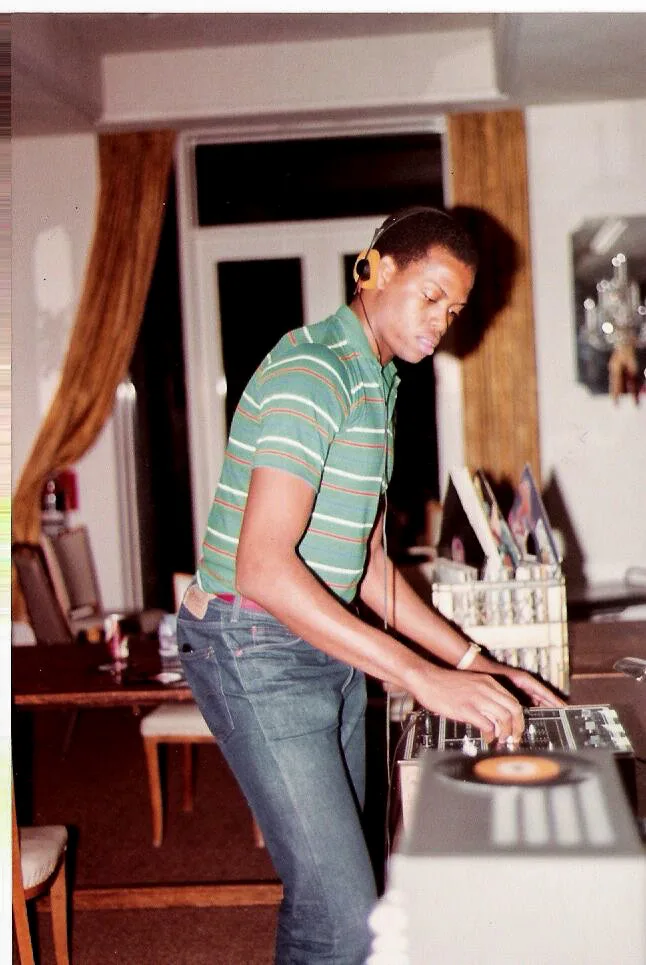 A teenage Craig Robinson plays with a DJ equipment. He has a buzz cut and is wearing black headphone with orange ear cups, and a green polo shirt with white, orange, and red stripes.