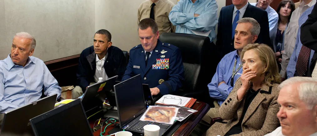 President Barack Obama wearing a navy blue jacket and a white polo shirt sits in a room with other individuals of a light skin tone. There is a brown table in front of them with four laptops and papers on it.