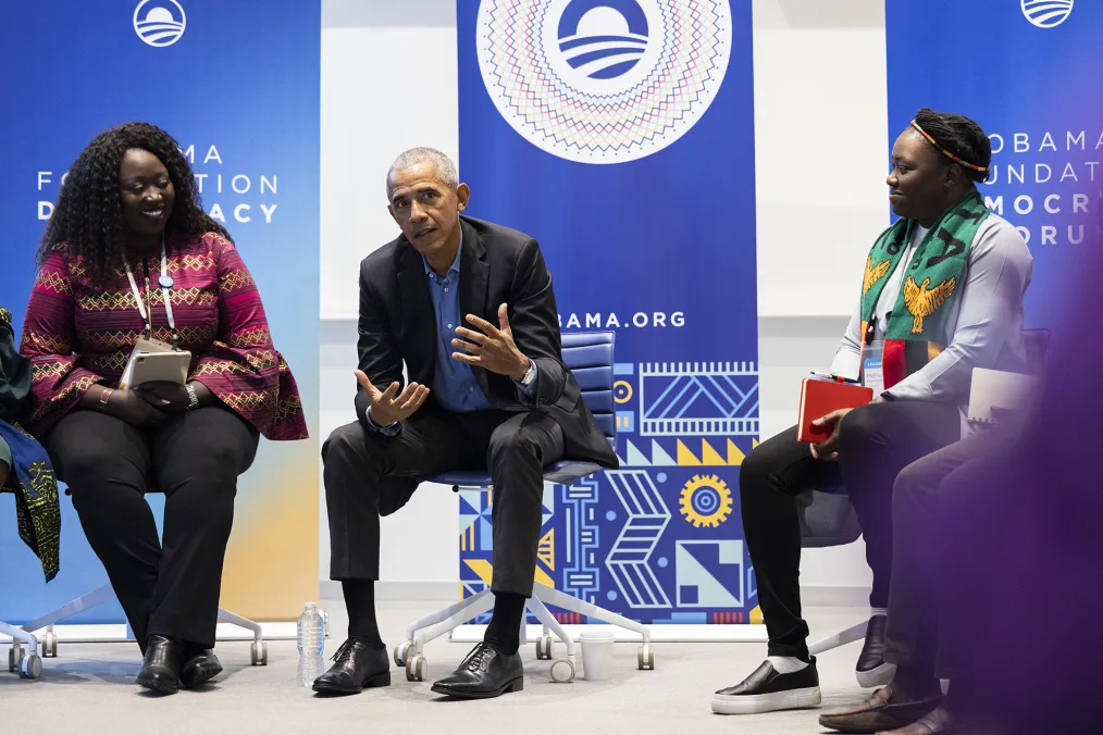 President Obama sits forward in a chair and speaks  with his hands gestured forward. He wears a dark suit and a blue button down shirt. Seated on either side of him are two young women with deep skin tones holding notebooks and wearing  colorful tops. Behind them, blue panels that read "Obama Foundation Democracy Forum" with the Obama Leaders: Africa logo. 