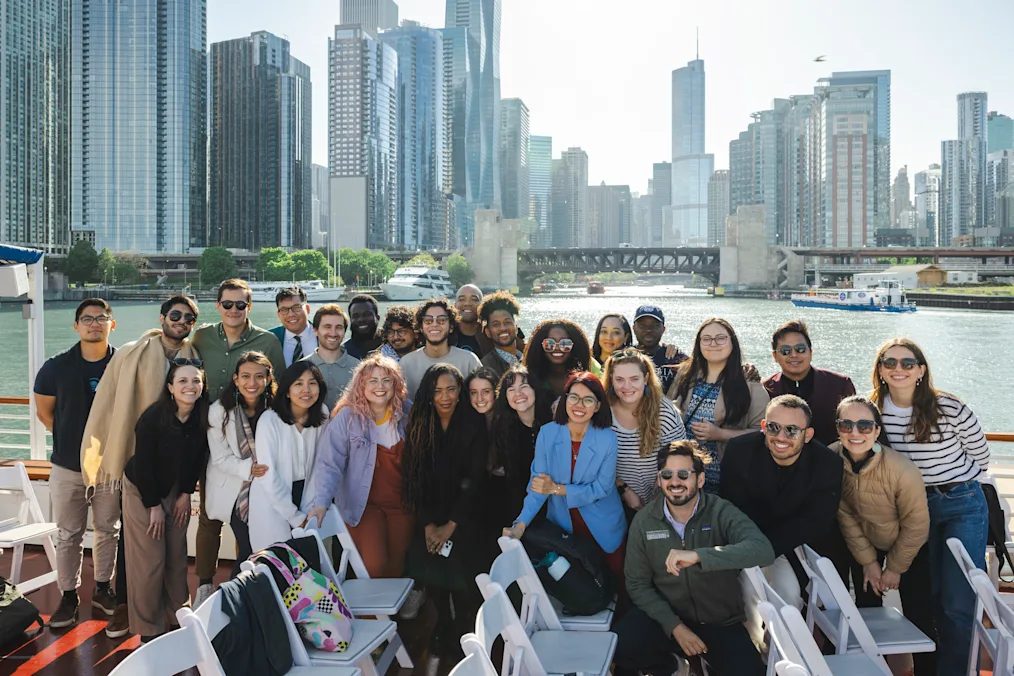 In front of the Chicago River, a group of people with a range of light to dark skin tones pose for a photo. There’s a mix of casual and professional clothing. 