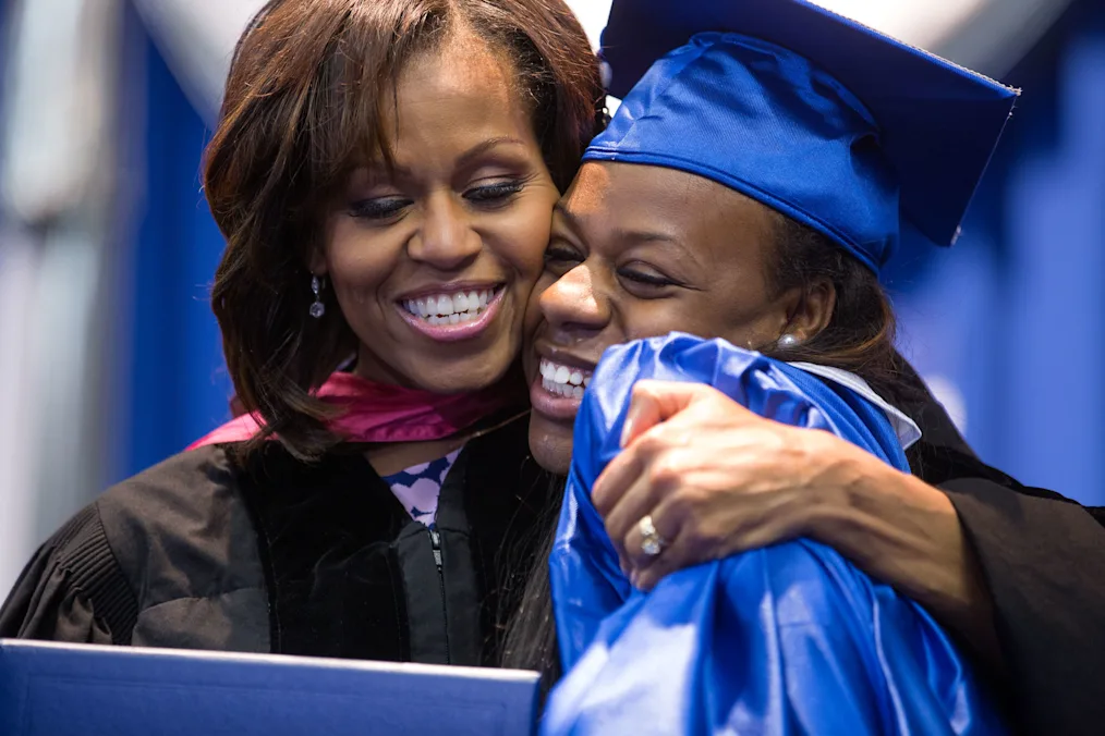 Mrs. Obama hands a diploma to a graduating senior, a  young Black woman with a dark skin tone wearing a royal blue gown, during the Martin Luther King, Jr. Academic Magnet High School for Health Sciences and Engineering at Historic Pearl High commencement ceremony.
