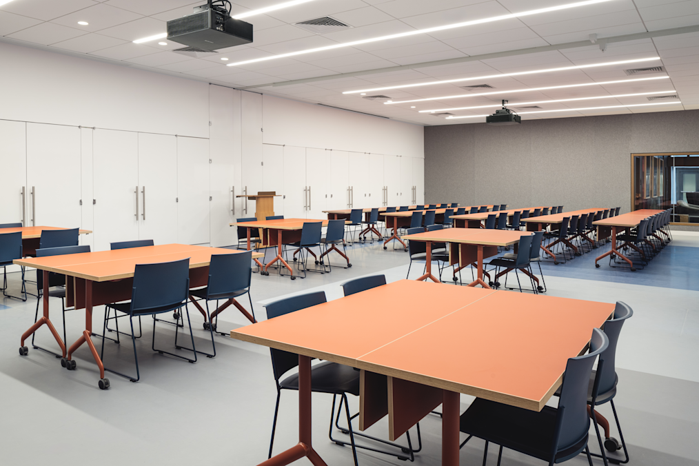 Orange-topped classroom tables with dark blue chairs arranged facing toward the front of a room with white paneled walls. In the foreground, a large rectangular table formed by two smaller orange tables pushed together has four dark blue chairs positioned around it.
