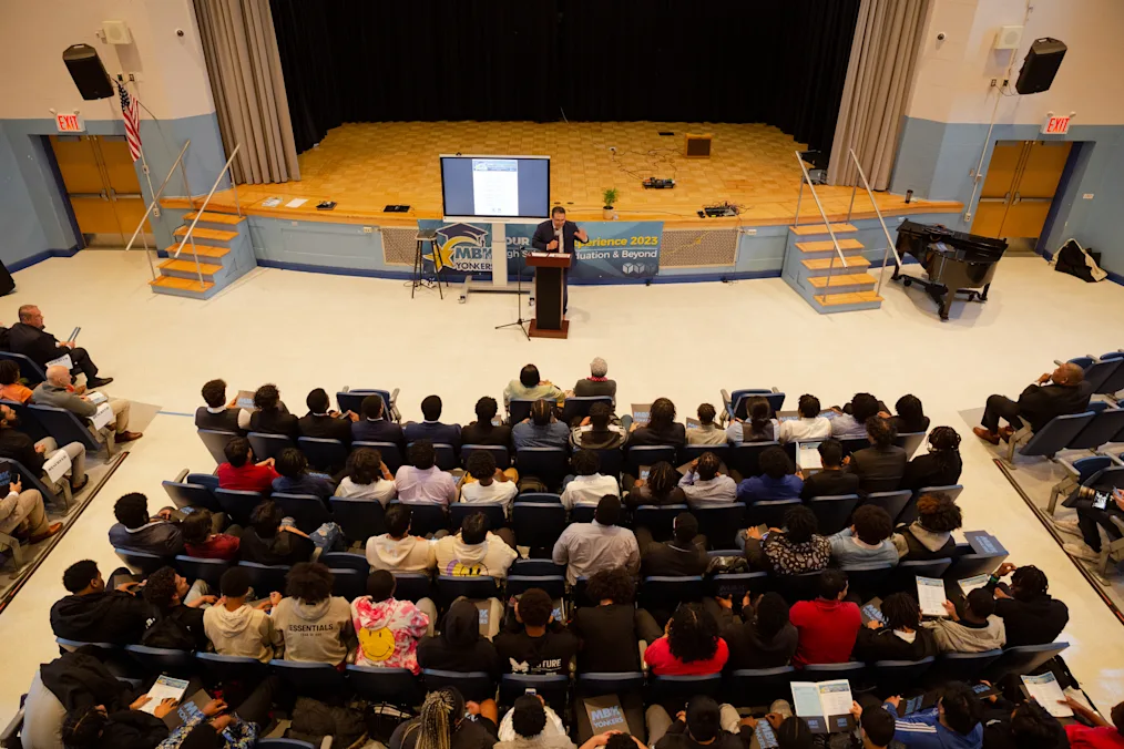 An aerial view of an audience with a range of people with light to dark skin tones. A man with a medium skin tone speaks behind a podium. He is wearing a blue suit jacket and glasses. 